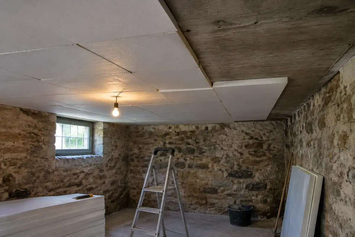 Panneaux isolants rigides fixés sous la dalle béton d'une cave aux murs en pierre, une partie du plafond encore nue